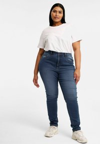 Höga blå denimjeans med slim fit, framfickor och klassisk knappstängning, i kombination med en vit t-shirt med kort ärm.
