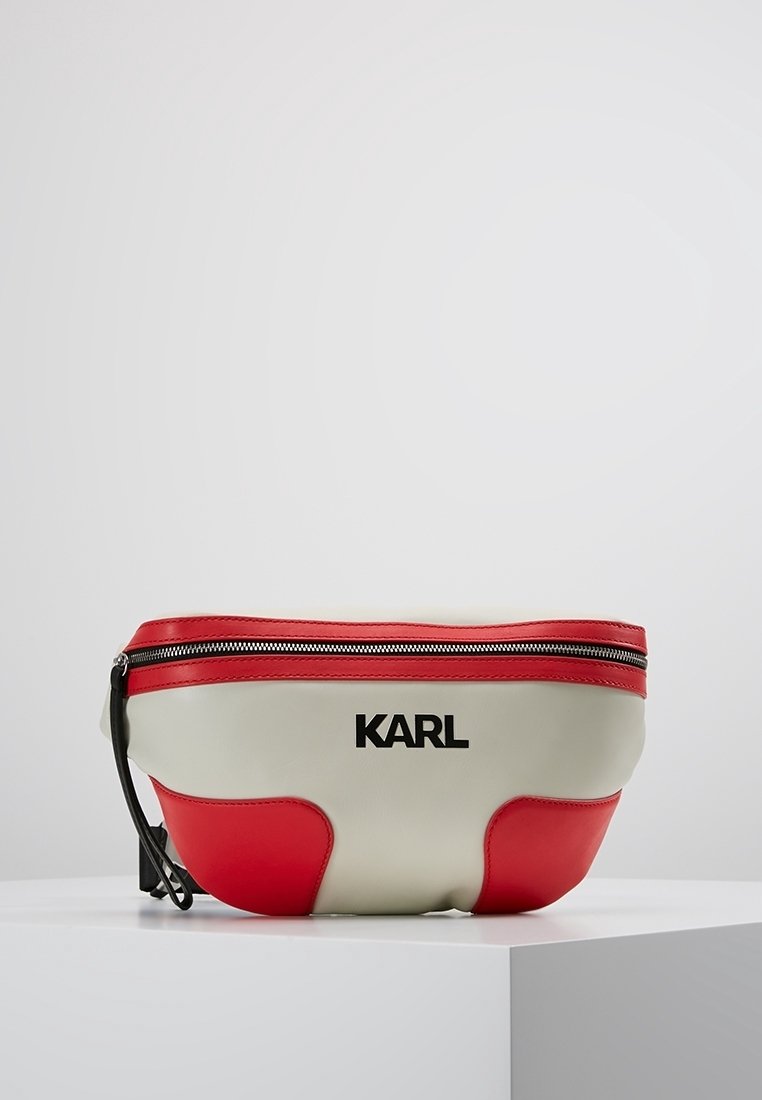 Sac banane en cuir blanc et rouge avec fermeture éclair et logo noir "KARL" centré sur le devant, placé sur un socle blanc sur fond gris.