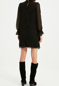 Schwarzes Kleid mit durchscheinenden langen Ärmeln, Rüschenkragen und Knopfverschluss hinten. Kombiniert mit knielangen schwarzen Wildlederstiefeln.