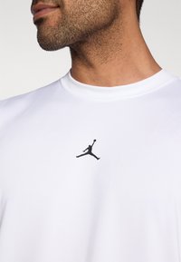 T-shirt de sport blanc avec un col rond, arborant un logo Jumpman noir centré sur la poitrine. Le tissu semble lisse et léger.