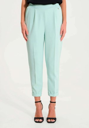 Lyse mintgrønne, tætsiddende bukser med opbuk ved buksebenene, båret med sorte, ankelstroppede højhælede sandaler mod en ensfarvet baggrund.