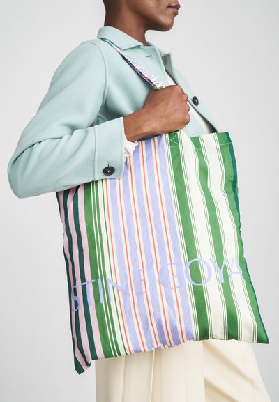 Randig tote-väska i flerfärgad tyg, med vertikala ränder i grönt, rosa, vitt och lavendel, samt varumärket "STINE GOYA" i ljusblått.