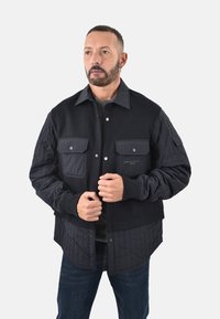 Giacca nera trapuntata con una combinazione di tessuto liscio e texturizzato, dotata di tasche frontali e bottoni a pressione, indossata sopra un maglione scuro.