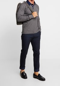 Polo gris à manches longues avec un col bleu marine, associé à un pantalon ajusté bleu foncé et des mocassins noirs, complété par un sac à dos gris.