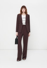 Costume marron foncé sur mesure avec blazer croisé et pantalon à jambes larges, associé à un t-shirt graphique blanc et un petit sac à main noir.