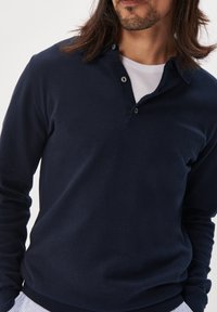 Marineblauer Strickpullover mit einem Knopfkragen, der eine weiche Textur und gerippte Bündchen aufweist. Der Stoff wirkt leicht und glatt.