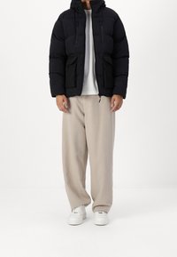 Giacca a vento nera con cappuccio, con due tasche frontali e chiusura con cerniera; indossata sopra una camicia bianca e pantaloni beige ampi.