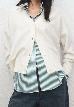 Personne portant un cardigan blanc boutonné par-dessus une chemise rayée verte et blanche avec un pantalon sombre, debout contre un mur uni.