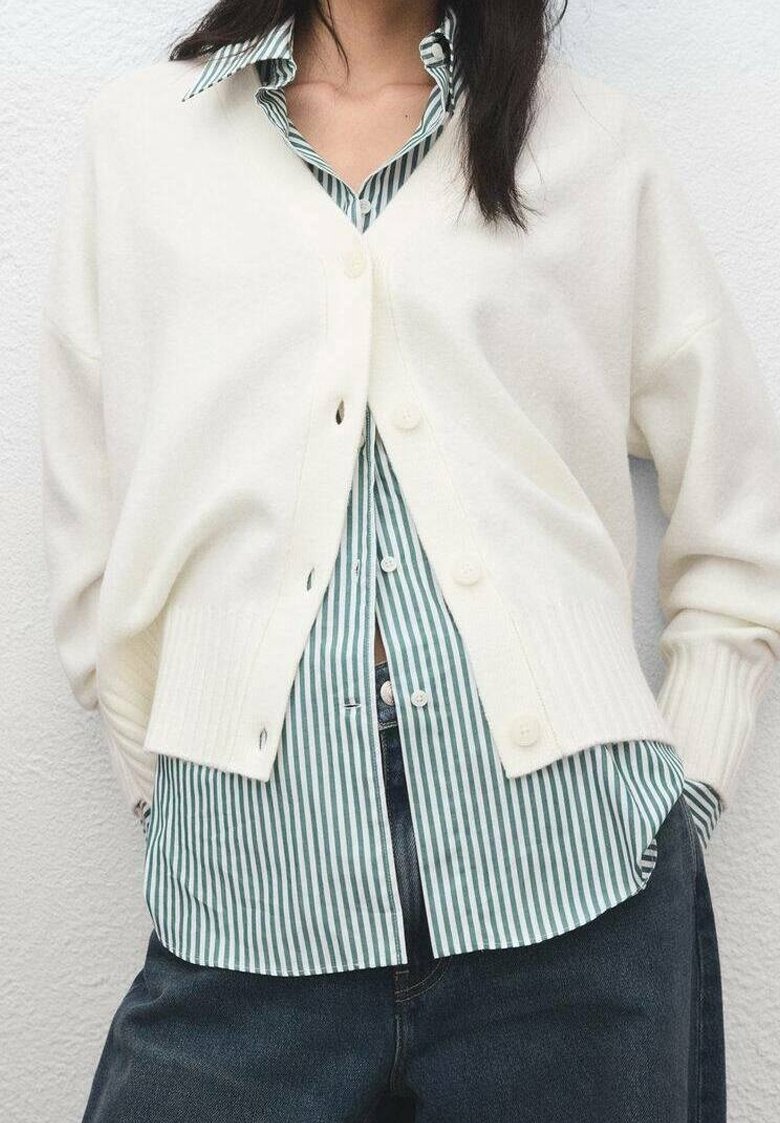 Personne portant un cardigan blanc boutonné par-dessus une chemise rayée verte et blanche avec un pantalon sombre, debout contre un mur uni.