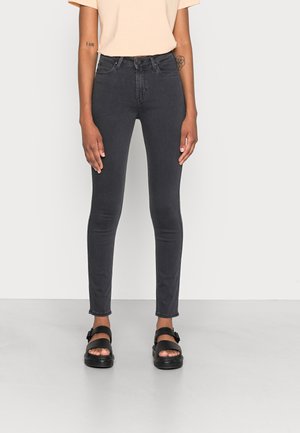 Persoon draagt zwarte nauwsluitende jeans, beige T-shirt met korte mouwen en zwarte sandalen met open tenen, staande voor een effen achtergrond.