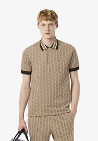 Beige pólóing, geometrikus mintával, fekete és arany csíkos gallérral és rövid fekete ujjakkal; Lacoste logó a mellkason.
