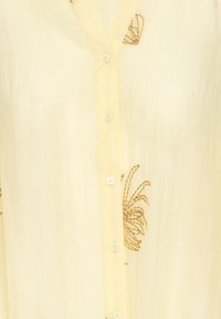 Tissu jaune clair avec boutons verticaux et motifs floraux brodés marron sur un matériau texturé.