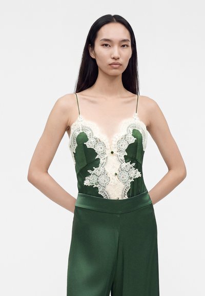 Jeune femme aux longs cheveux foncés portant un caraco en satin vert foncé avec des détails en dentelle blanche et un pantalon taille haute assorti sur fond blanc.