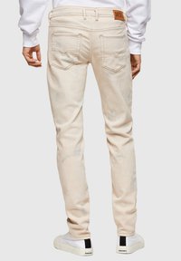Jeans de color crema con un ajuste slim, que cuentan con bolsillos traseros y un parche de cuero en la cinturilla. Texturizados con sutiles detalles desgastados.