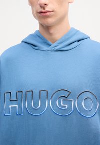 Felpa blu con un grande e audace logo "HUGO" ricamato in gradiente. Tessuto morbido con una vestibilità rilassata e cappuccio con cordino.