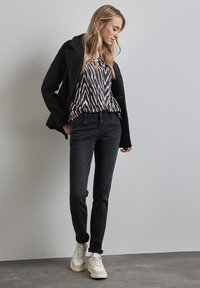 Czarne dżinsy o dopasowanym kroju, w połączeniu z wzorzystą bluzką w czarno-białe desenie oraz czarnym, teksturowanym płaszczem. Białe trampki dopełniają stylizację.