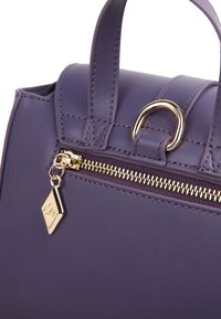 Borsa a tracolla in pelle viola con una cerniera dorata, accenti circolari e manici in tessuto coordinato; presenta un'etichetta decorativa con il logo.