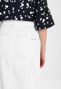 Lado derecho de una persona que lleva una falda de mezclilla blanca con un bolsillo trasero y una blusa azul marino con lunares blancos y mangas acampanadas.