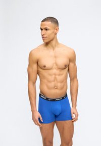 Μυώδης άνδρας όρθιος, χωρίς πουκάμισο, φορά μπλε μπόξερ της Nike με μαύρη ζώνη, κοιτώντας ελαφρώς προς τα αριστερά πάνω σε απλό φόντο.