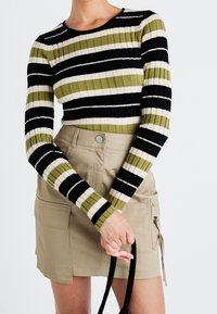Gestreept ribgebreid top in zwart, groen en crème, gecombineerd met een beige utilitaire rok met zakken en koordaccenten.