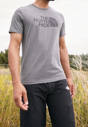 Mann trägt ein graues The North Face T-Shirt und schwarze Shorts steht draußen auf einer Graswiese.