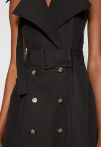 Trench-coat noir avec un design à double boutonnage, un col large et une ceinture ajustable. Tissu texturé avec des boutons brillants en accents.