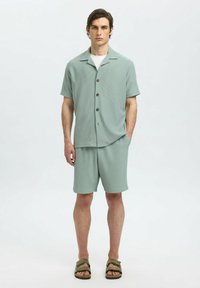 Camicetta verde chiaro a maniche corte con bottoni e un motivo texturizzato, abbinata a pantaloni corti coordinati. Il modello indossa sandali marroni a ciabatta.