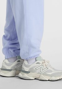 Jalat, joissa on valkoiset ja harmaat New Balance -lenkkarit sekä valkoiset sukat, yhdistettynä vaaleanpunaiseen jogger-housuihin yksinkertaista taustaa vasten.