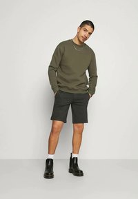 Pantalones cortos gris oscuro con bolsillos laterales, combinados con una sudadera verde oliva. Botines negros hasta el tobillo y un detalle de cadena plateada completan el atuendo.