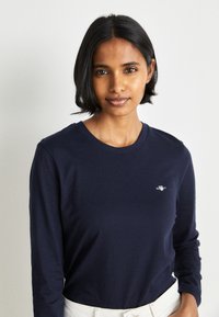 Marineblå langermet t-skjorte laget av bomull, med rund hals og en liten logo på venstre bryst. Myk tekstur og tettsittende design.