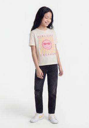 Κορίτσι με μακριά σκούρα μαλλιά φοράει λευκό t-shirt με τη φράση «Paradise Dreamer», σκισμένα μαύρα τζιν και λευκά αθλητικά παπούτσια με κίτρινο σήμα, στέκεται και χαμογελάει.