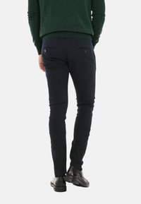 Pantaloni slim-fit blu scuro con tasche posteriori, realizzati in un morbido misto cotone, abbinati a scarpe di pelle nere e un maglione verde.