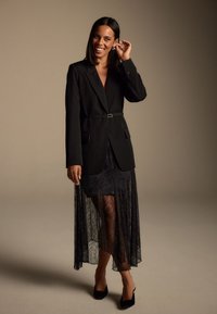 Black blazer with belt detail over a sheer black lace skirt. Square-toed black heels complete the outfit. Simple background in neutral tones.