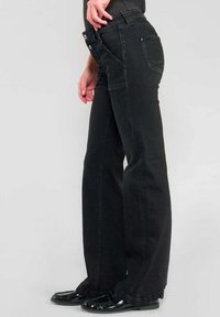 Vaqueros de mezclilla negros de pierna ancha con cintura alta, que cuentan con un cierre de botones, bolsillos delanteros y un bolsillo trasero, llevados con zapatos negros brillantes.