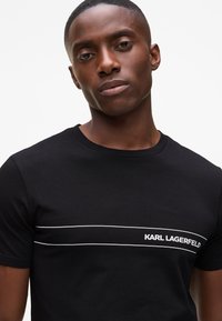 Fekete pamut póló rövid ujjakkal, fehér vízszintes csíkkal, és az "KARL LAGERFELD" felirattal hímezve az alsó jobb oldalon.