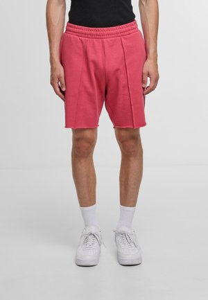 Rosa Sportshorts mit Gummibund, gefältetem Vorderdesign und ausgefranstem Saum. Getragen mit weißen Turnschuhen und Socken.