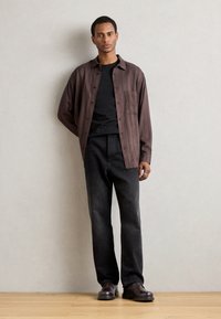 Camicia a righe marroni con bottoni sopra una maglietta nera, abbinata a jeans neri a gamba dritta e scarpe marroni, in piedi su uno sfondo semplice.