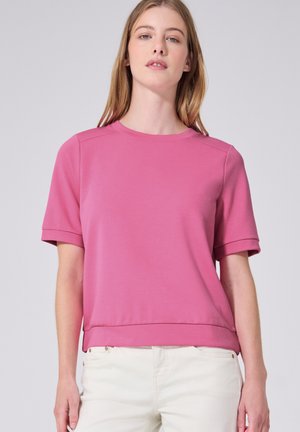 Joven mujer con cabello rubio lacio, vistiendo una camiseta de manga corta rosa y pantalones blanco hueso sobre un fondo gris claro.