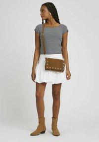 Sac à bandoulière en daim marron avec des clous argentés et une chaîne, porté avec un haut rayé noir et blanc et une jupe évasée blanche.