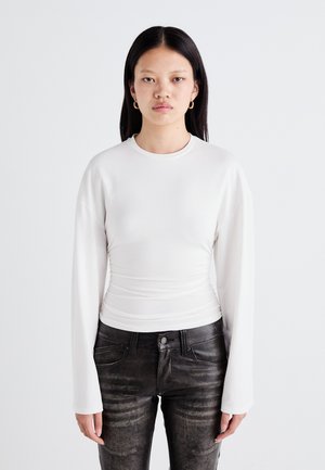 Hvit langermet topp med en tilpasset silhuett og rynket tekstur ved midjen, kombinert med svarte utslitte jeans.
