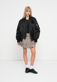 Veste bomber noire avec une texture lisse, coupe oversize et poche zippée sur le côté, associée à une jupe à imprimé léopard et des baskets noires.