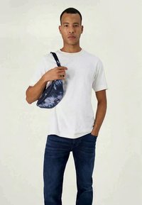 Uma pochete preta e azul tie-dye usada sobre uma t-shirt branca lisa. O modelo está de jeans azuis, com as mãos posicionadas de forma casual.