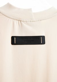 Camisa de tela color crema con cuello redondo que presenta una etiqueta rectangular negra cosida cerca del cuello con texto en relieve.