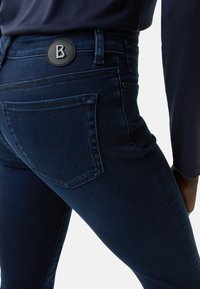 Tmavě modré skinny džíny vyrobené z denimu, s kulatou černou nášivkou s bílým logem "B" na zadní straně a jednou viditelnou zadní kapsou.