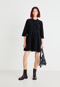 Rochie neagră cu nasturi, cu un model texturat, mâneci trei sferturi și talie strânsă. Îmbrăcată cu botine negre și roșii până la gleznă și o geantă neagră.
