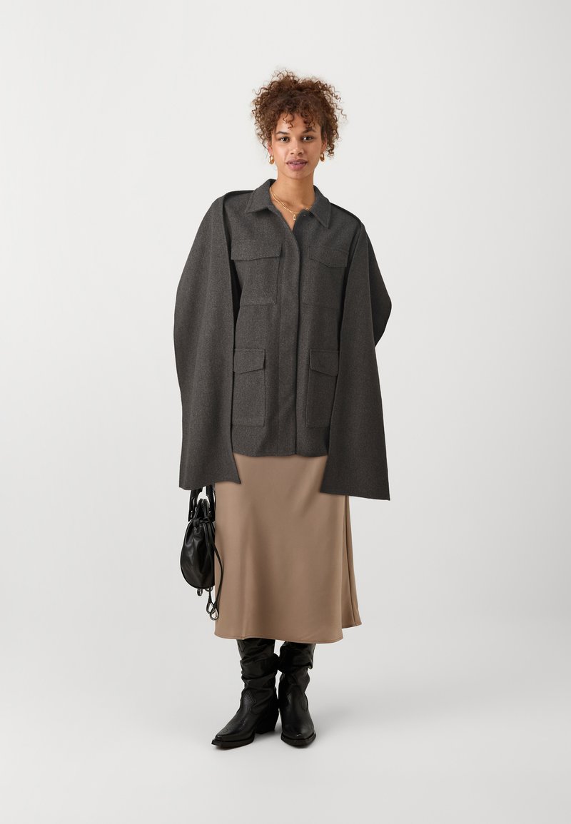 Grijze wolmix overhemdjas met oversized mouwen en voorzakken, gecombineerd met een beige middellange rok en zwart kniehoge laarzen.