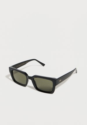 Gafas de sol rectangulares negras con lentes verde oscuro y montura de acabado mate, descansando sobre una superficie blanca.