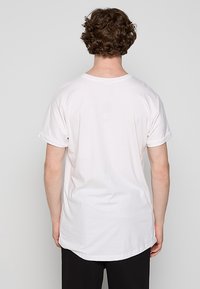 Homme aux cheveux bouclés portant un t-shirt blanc uni à manches courtes et un pantalon noir, vu de dos sur un fond gris uni.