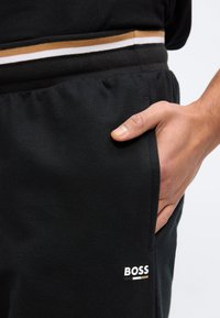 Κοντινό πλάνο χεριού που βρίσκεται στην τσέπη μαύρων παντελονιών φόρμας με το λογότυπο BOSS και ριγωτή μέση σε μαύρο τοπ.