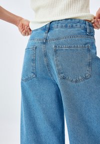 Vaqueros de mezclilla azul de tiro alto con un corte relajado, que presentan dos bolsillos traseros y un acabado deslavado. La tela parece resistente con costuras visibles.